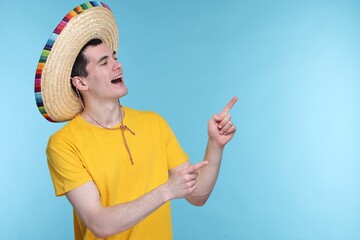 Young man in Mexican sombrero hat pointing at something on light blue background. Space for text