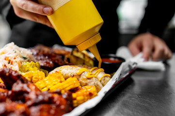 A close-up of someone drizzling mustard onto grilled corn, served alongside BBQ ribs and coleslaw on a tray. The ribs are well-cooked, and the background appears to be a kitchen or restaurant