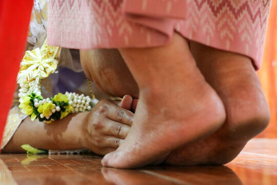 A young Thai man who is about to be ordained as a Buddhist monk clasps his hands together and bows down to the feet of his biological mother in gratitude.