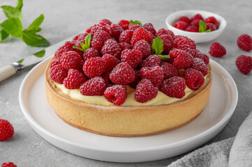 Delicious raspberry tart with vanilla custard cream sprinkled with powdered sugar on a white plate on a gray concrete background. Summer dessert.