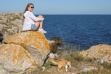 mature woman with long blond hair sits on a stone on the seashore, a cat on a leash walks nearby. Traveling and tourism with pets