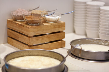 In the kitchen, two pots of oatmeal are cooking. On the wooden crate, three glass bowls hold cocoa, sugar, and grated chocolate, each with a metal spoon. Neat stacks of white plates are placed nearby.