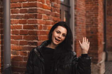Happy attractive woman in black bomber, walk outdoors, meeting her friends or love, showing high five hey hello hi sign. She is joyful to meet her acquainted by chance. Girl wave her hand.