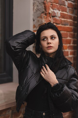 Trendy girl with brunette hairstyle posing outside. Woman in leather puffer jacket fix hair,...