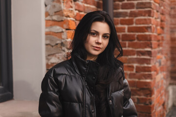 Beautiful woman smiling near brick wall. Outdoor portrait of a smiling brunette girl. Happy cheerful girl look sensual, dreaming at city, look fashion. She wear black puffer jacket.