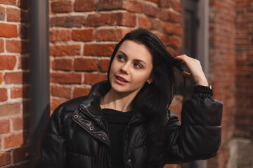 Trendy girl with brunette hairstyle posing outside. Woman in leather puffer jacket fix hair, walking on the street outdoors. Tourist happy woman posing in the city. Optimistic lady walks outside.