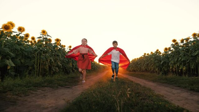 Happy children superhero in red cloak running image flying at sunset sunflower field. Boy and girl playing hero flight for protect planet fantasy imagination at sunrise sun light agriculture meadow