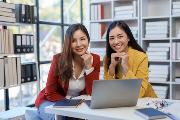 Two pretty young Asian businesswoman sitting at desk with laptop doing paperwork together discussing project financial report. Corporate business collaboration concept.