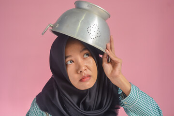 An Asian woman wearing a green shirt and black hijab is wearing a pot on her head. Cooking theme