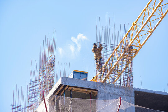Construction worker high up on a building frame fixing rebar mesh next to a yellow crane jib under a bright blue sky.