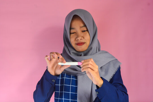 Happy and grateful young woman wearing blue shirt with gray hijab showing her pregnancy test, hand on chest smiling isolated on pink background
