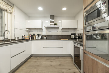 Frontal image of a furnished kitchen in a single-family home with integrated stainless steel appliances and several windows on one side