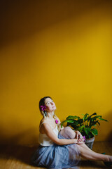 Portrait of a blonde girl against a yellow wall. The girl is sitting on the floor next to a flower in a pot