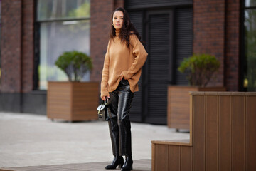 Beautiful fashion model in the latest trends layered knit sweater, leather pants, high heeled boots Standing on a city street