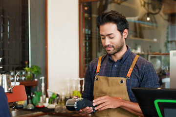 Asian male cafe worker in a plaid shirt examines handheld payment device with focused attention. Barista in work apron looks at electronic payment terminal, concentration evident in well-lit eatery