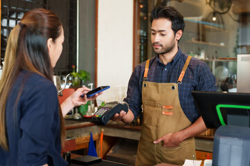 Asian barista in a checkered shirt engages with customer, processing payment on a portable device...