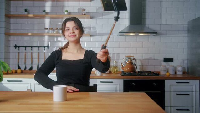 Beautiful young woman sitting in the kitchen smiling and taking bunch of selfie photos with selfie stick