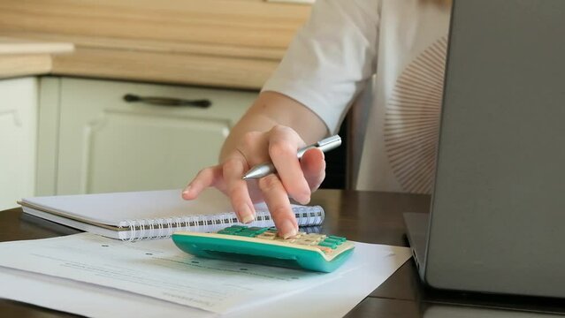 A woman holds a pen and using calculator in her home office. Desktop with a note, laptop, pen and documents. HD Video footage