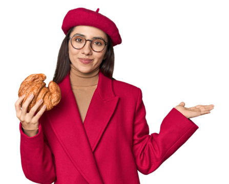 Chic Caucasian woman eating croissant in red coat and beret on studio background showing a copy space on a palm and holding another hand on waist.