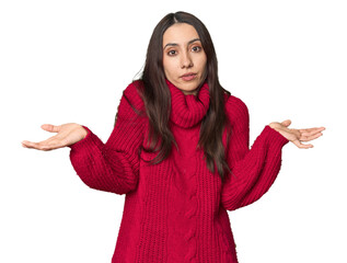 Young Caucasian woman in cozy red sweater on studio background doubting and shrugging shoulders in...