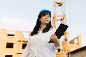 Plus size female engineer in a white shirt and blue helmet looking up. 
Female in blue helmet architect is holding papers at a construction site. 