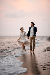 Happy smiling newlyweds walk on the water along the seashore, holding hands