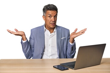 Elegant businessman at desk with laptop confused and doubtful shrugging shoulders to hold a copy space.