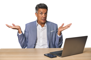 Elegant businessman at desk with laptop confused and doubtful shrugging shoulders to hold a copy space.