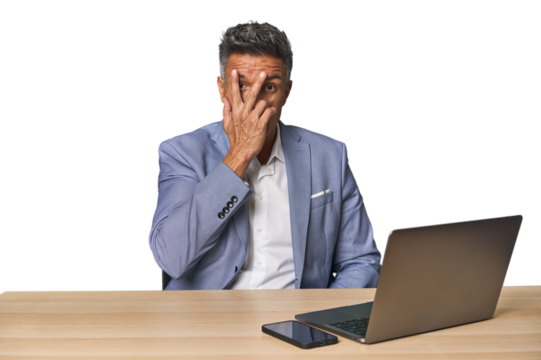 Elegant businessman at desk with laptop blink at the camera through fingers, embarrassed covering face.