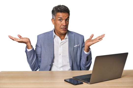 Elegant businessman at desk with laptop confused and doubtful shrugging shoulders to hold a copy space.