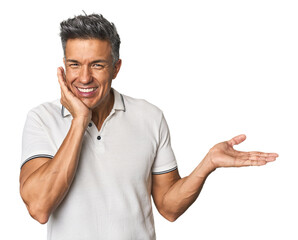 Middle-aged Latino man holds copy space on a palm, keep hand over cheek. Amazed and delighted.