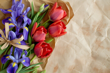 A composition of red tulips and purple irises, wrapped in a brown paper and tied with a ribbon.