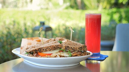 Outdoor brunch scene, fresh club sandwich on plate with side snacks and tall glass of red juice, served on table with natural backdrop. Casual dining and healthy eating.