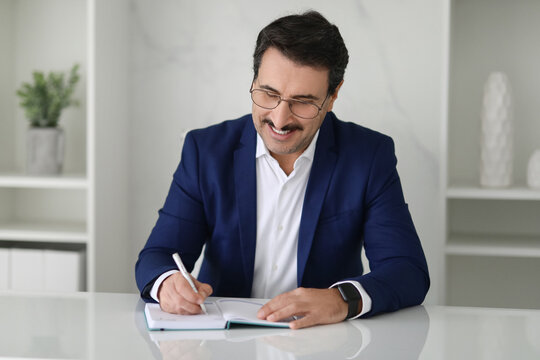 Content businessman in a blue suit writing in a notebook with a smile - Powered by Adobe