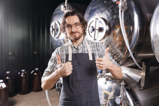 Portrait happy young bearded man shows thumbs up on background craft beer tank. Brewery worker with industrial equipment - Powered by Adobe