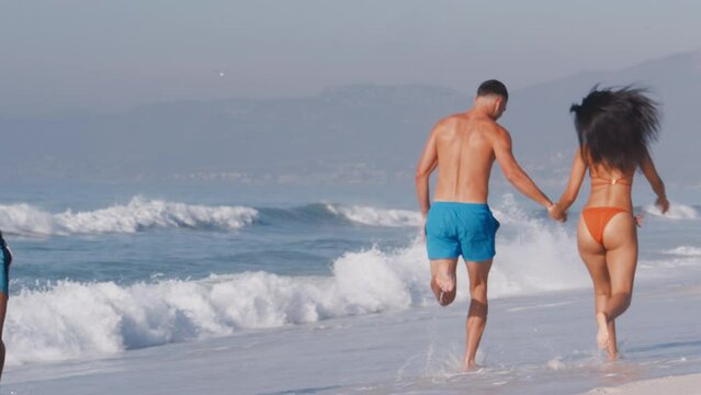 Rear view of group of young couples in swimwear having fun on summer vacation holding hands and running along beach in South Africa - shot in slow motion