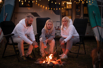 A family cooks sausages on a bonfire near their motorhome in the woods