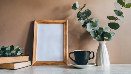 empty wooden picture frame poster mockup hanging on beige wall background vase with green eucalyptus tree branches on table cup of coffee books working space home office