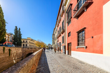 Pedestrians at the entrance to the narrow, historic Carrera del Darro street of shops, medieval buildings and the Darro river in the picturesque center of the Andalusian city of Granada, Spain