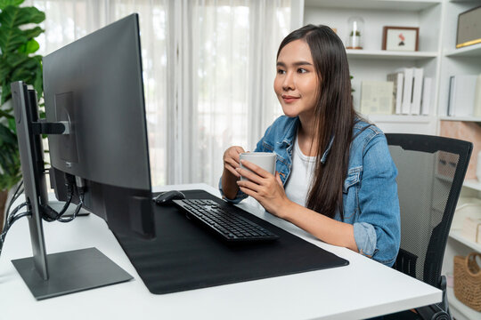 Working young beautiful asian creative manager with glasses focusing on pc while drinking hot coffee, creating with website design on social media with blue jeans shirt at home office. Stratagem.