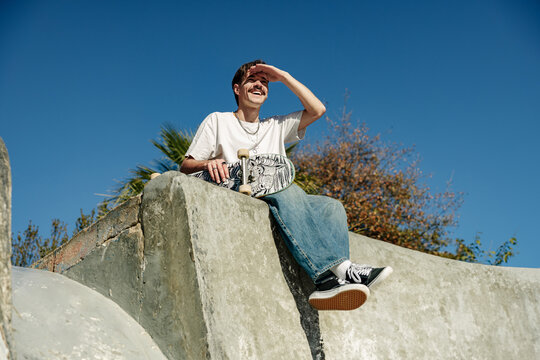 Low angle of young male skateboarder holding skateboard sitting on ramp in skate park  - Powered by Adobe