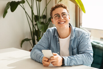Cheerful young man with curly hair and glasses holding a smartphone and smiling