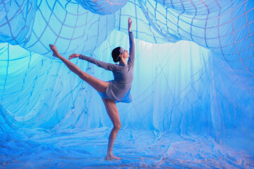 Ballerina dancer in a room with white drapes made of thin airy fabric, illuminated by blue spotlights.