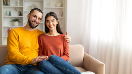 Loving man and woman sitting closely on sofa and smiling at camera
