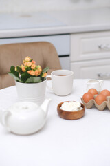 Fototapeta na wymiar Spring breakfast concept. White pot with flower, eggs, teapot, glass of tea and bowl with cottage on white table in light kitchen.