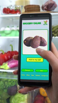 Vertically Oriented Image. Ordering Food Online Using Smartphone App. Close-up Of females Hands Marking Shopping List On Mobile Phone In Front Open Fridge. Food Delivery Mobile Application.