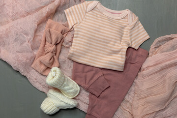 Flat lay of pink baby girl onesie and pants with headband and booties on pink scarf and gray background with copy space for gender reveal or baby girl announcement