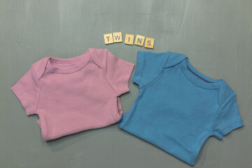Flat lay of pink girl and blue boy onesies on gray background with the letters Twins above for gender reveal or baby announcement