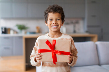 Happy little black boy proudly holding wrapped gift with bright red ribbon