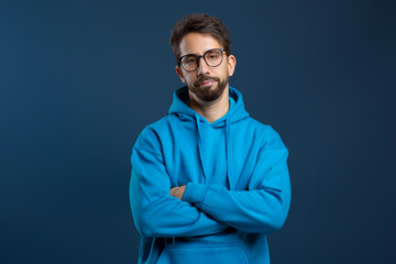 Man wearing glasses and blue hoodie looking disinterested, standing with arms crossed